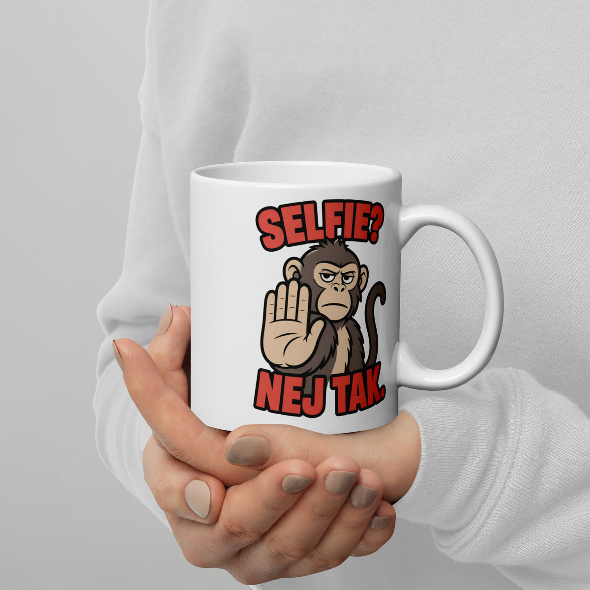 Hvidt keramikkrus med teksten Selfie? Nej tak og en abe der holder hånden op, holdt i hænderne, hank til højre