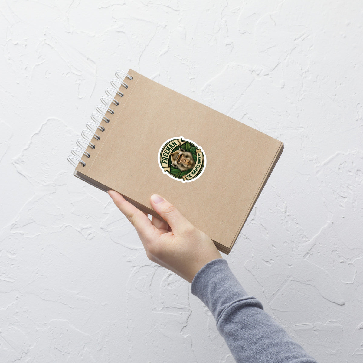 Klistermærke med Freeman The Miracle Monkey badge med en abe i grønt, 7,5 x 7,5 cm, på en notesbog