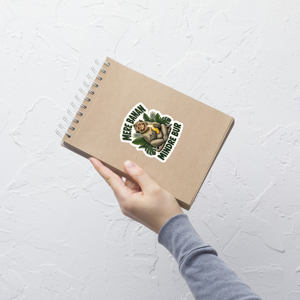 Klistermærke med teksten Mere banan, mindre bur og en glad abe med en banan, 10 x 10 cm, på en notesbog