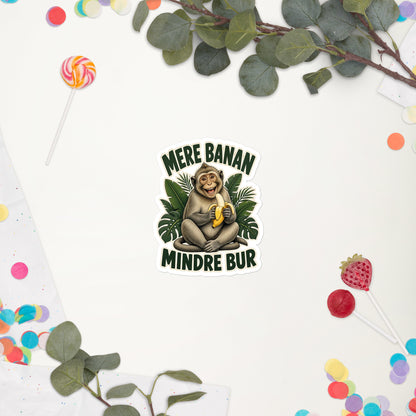 Klistermærke med teksten Mere banan, mindre bur og en glad abe med en banan, 10 x 10 cm, på en hvid baggrund med blomster