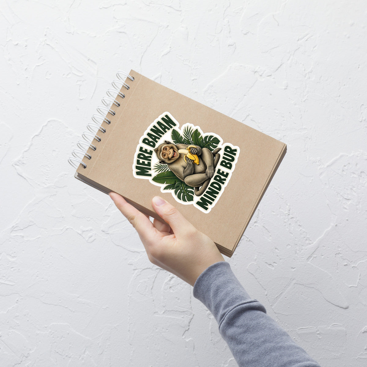 Klistermærke med teksten Mere banan, mindre bur og en glad abe med en banan, 14 x 14 cm, på en notesbog
