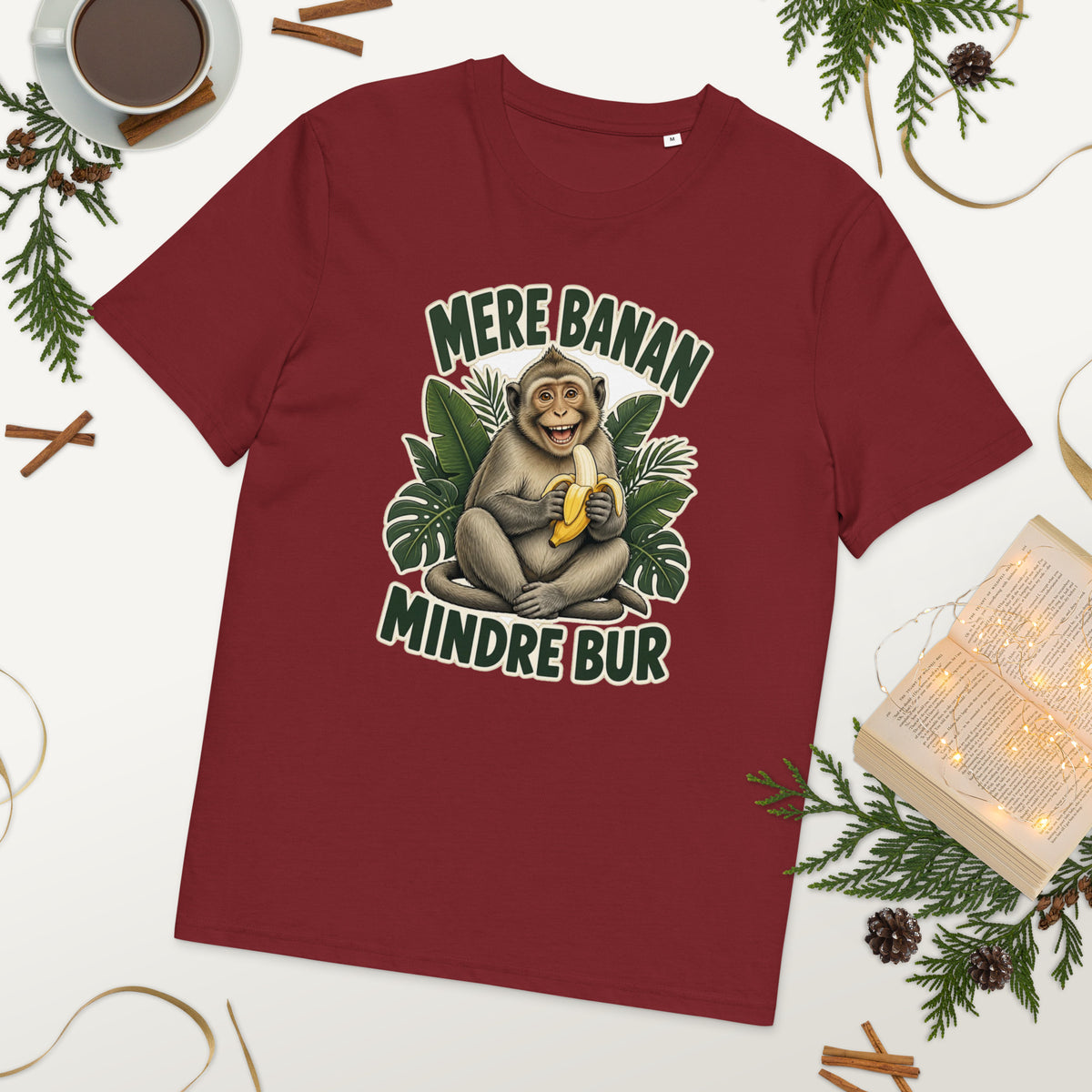 Abe t-shirt med teksten Mere banan, mindre bur i farven bordeaux, lagt på et bord med kaffe og grønt
