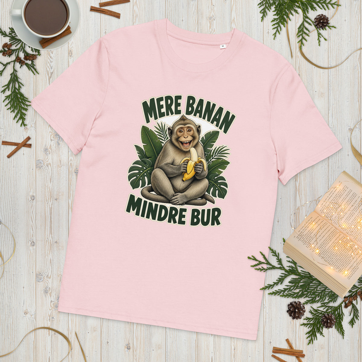 Abe t-shirt med teksten Mere banan, mindre bur i farven lyserød, lagt på et lyst træbord