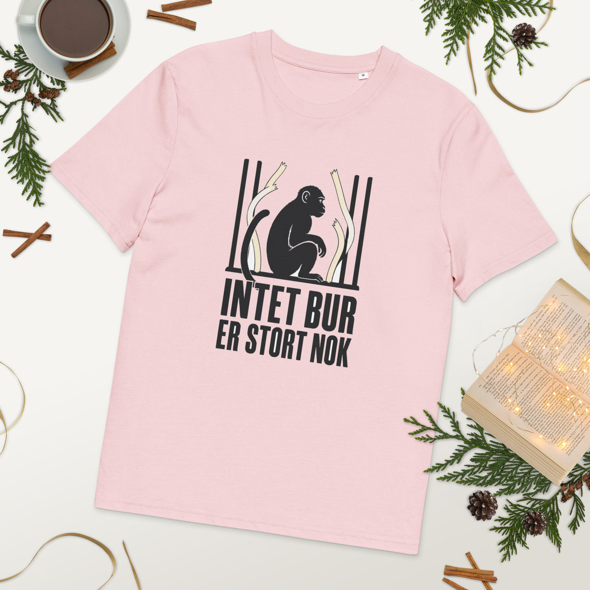 T-shirt med teksten Intet bur er stort nok i farven lyserød, lagt på et bord med kaffe og grønt