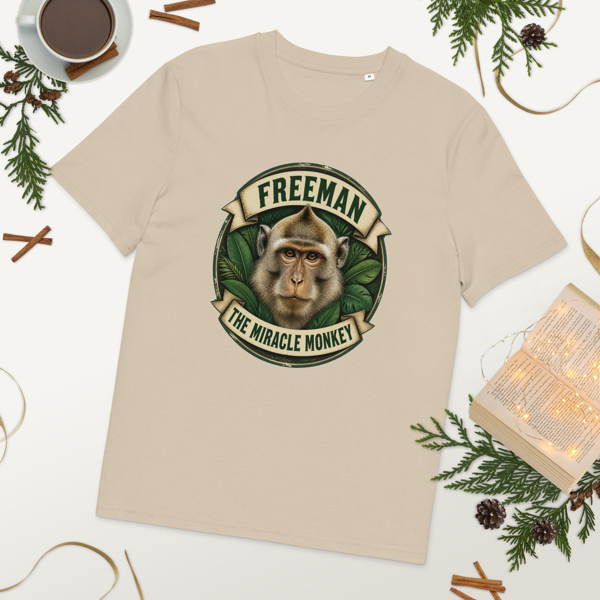 Freeman The Miracle Monkey t-shirt med abe i grønt badge i farven sand, lagt på et bord med kaffe og grønt
