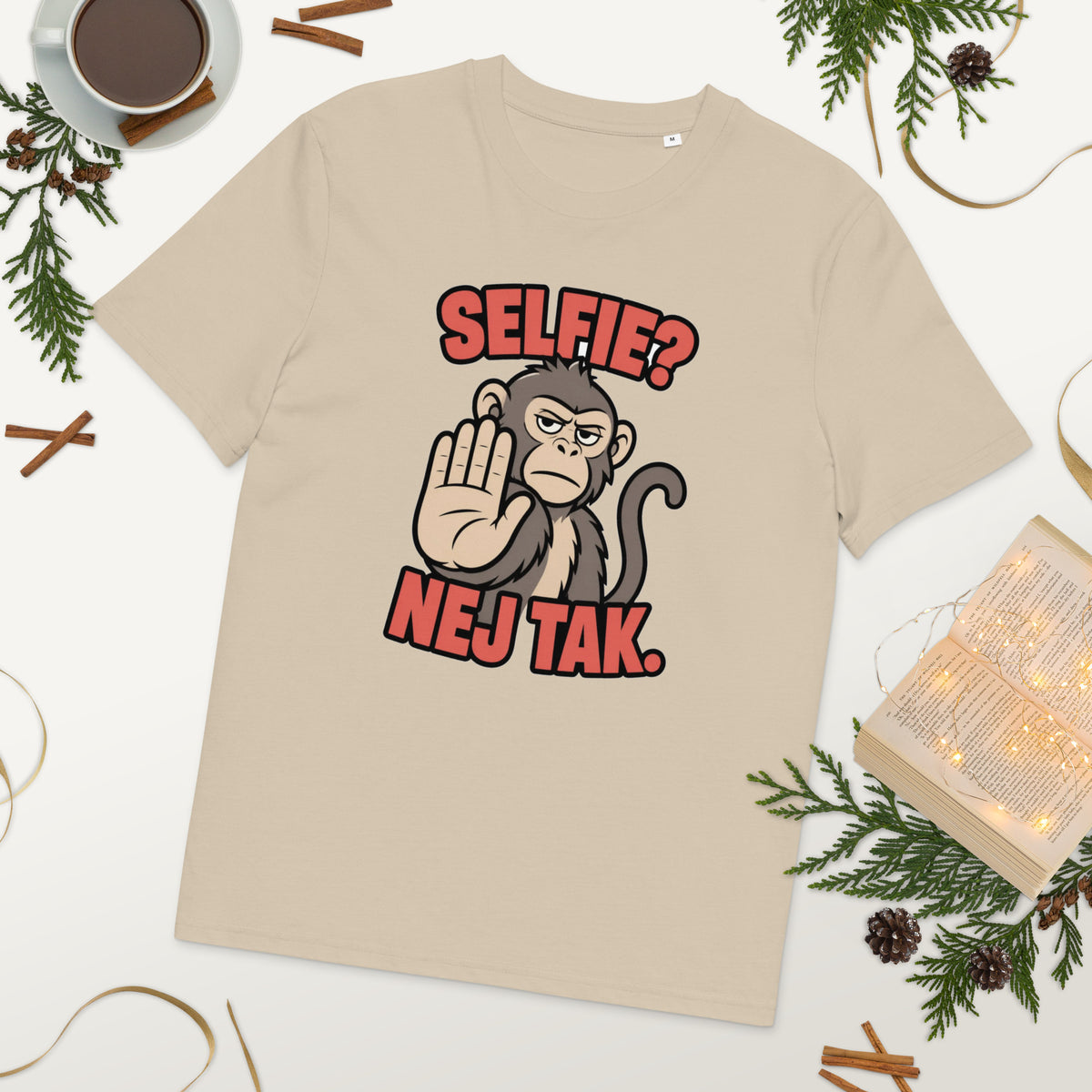 Sjov abe t-shirt med teksten Selfie? Nej tak. i farven sand, lagt på et bord med kaffe og grønt