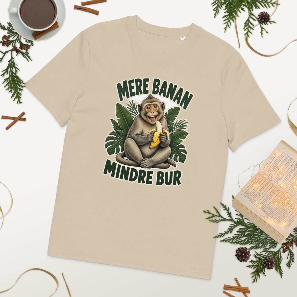 Abe t-shirt med teksten Mere banan, mindre bur i farven sand, lagt på et bord med kaffe og grønt
