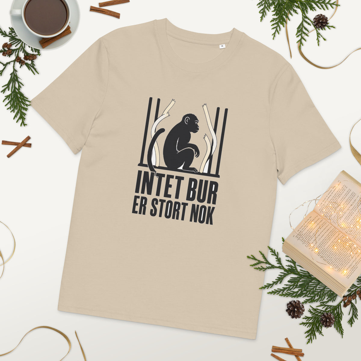 T-shirt med teksten Intet bur er stort nok i farven sand, lagt på et bord med kaffe og grønt