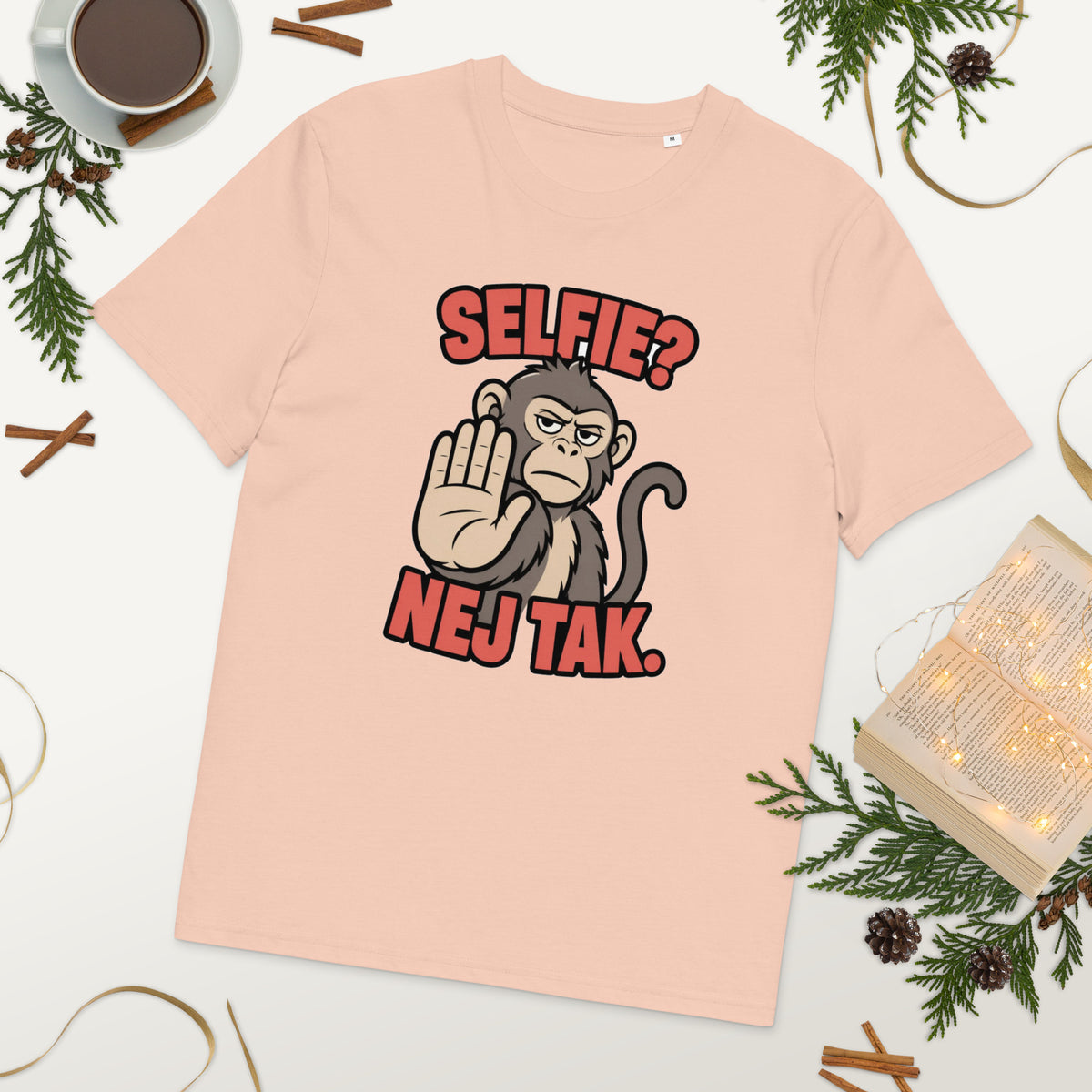 Sjov abe t-shirt med teksten Selfie? Nej tak. i farven fersken, lagt på et bord med kaffe og grønt