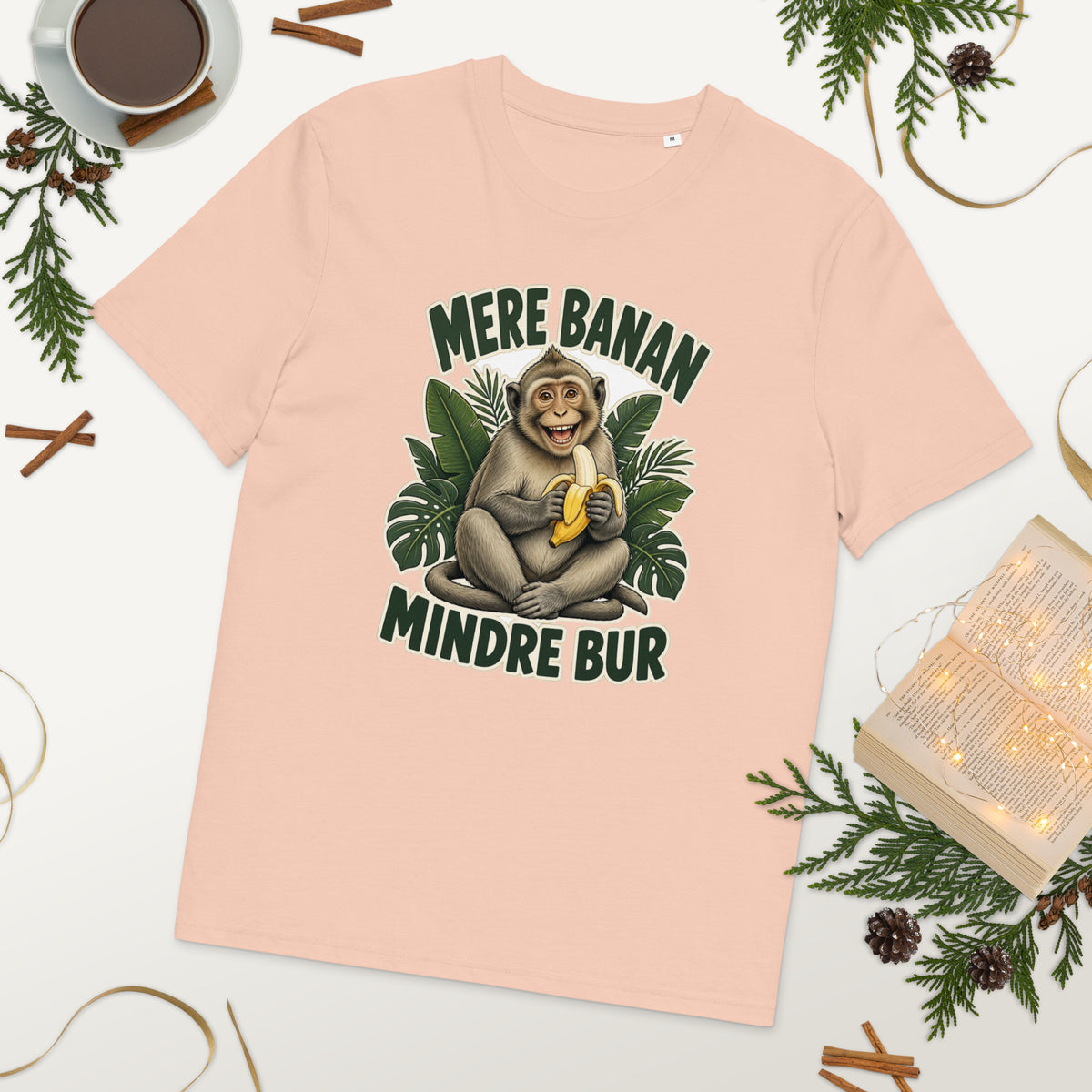 Abe t-shirt med teksten Mere banan, mindre bur i farven fersken, lagt på et bord med kaffe og grønt