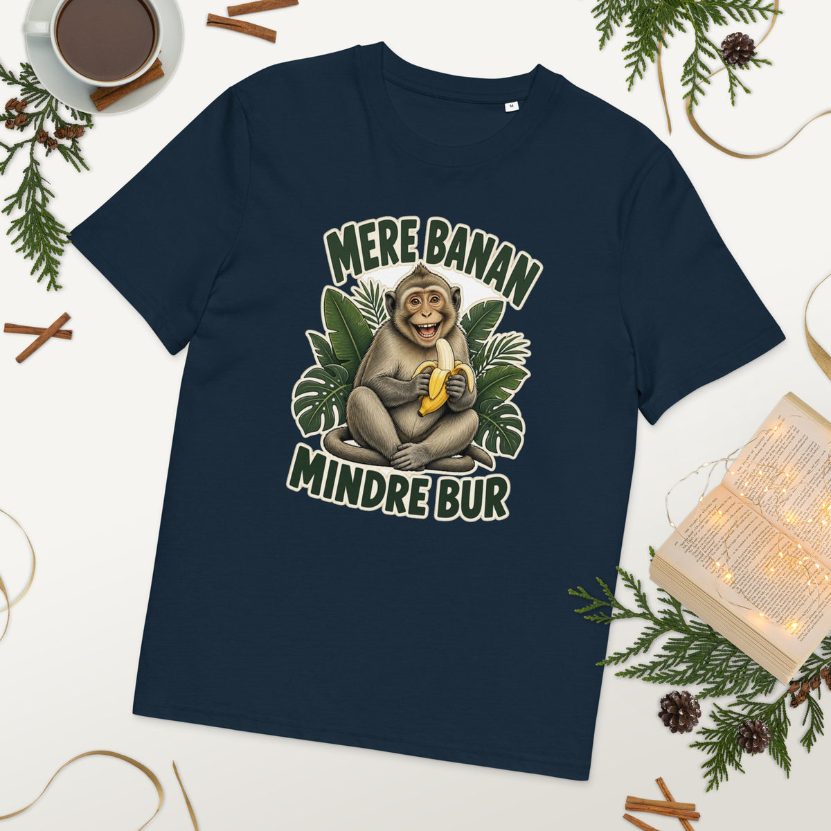 Abe t-shirt med teksten Mere banan, mindre bur i farven marineblå, lagt på et bord med kaffe og grønt