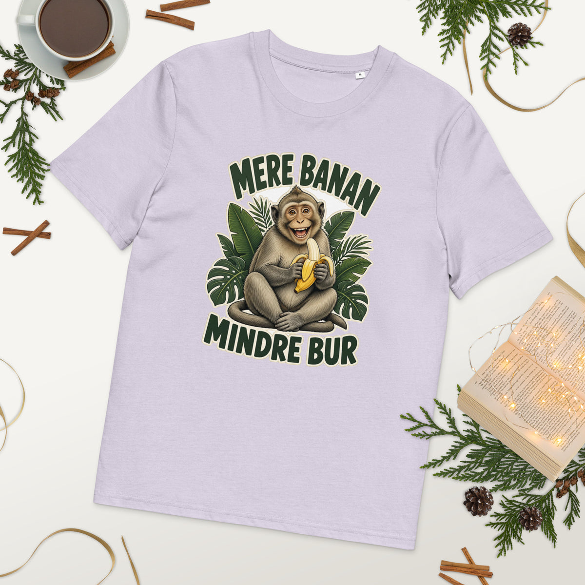 Abe t-shirt med teksten Mere banan, mindre bur i farven lavendel, lagt på et bord med kaffe og grønt