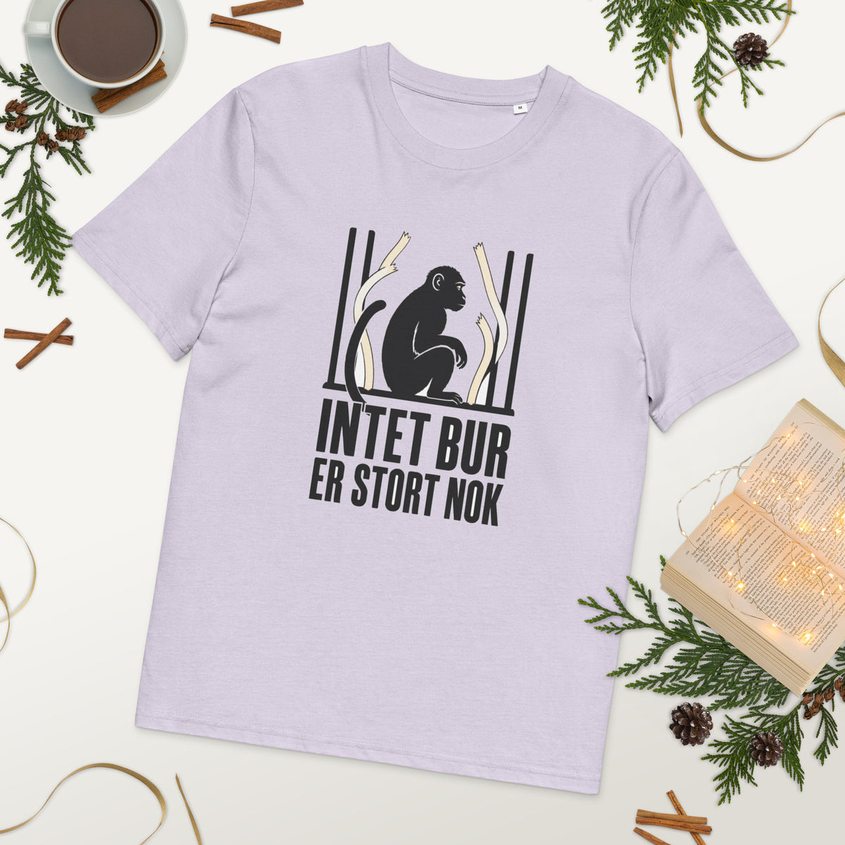 T-shirt med teksten Intet bur er stort nok i farven lavendel, lagt på et bord med kaffe og grønt