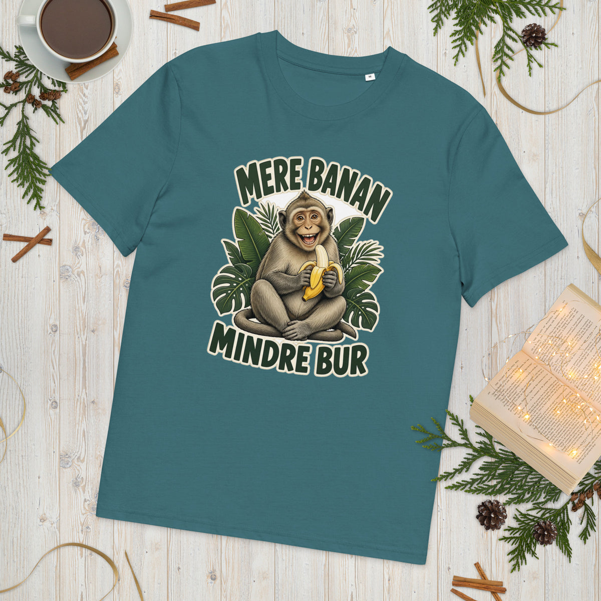 Abe t-shirt med teksten Mere banan, mindre bur i farven petroleumsblå, lagt på et lyst træbord