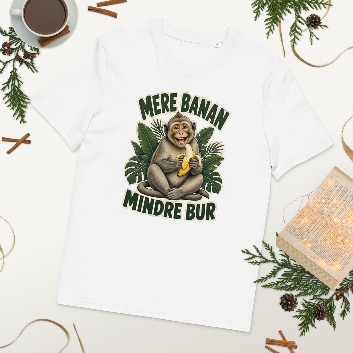 Abe t-shirt med teksten Mere banan, mindre bur i farven hvid, lagt på et bord med kaffe og grønt