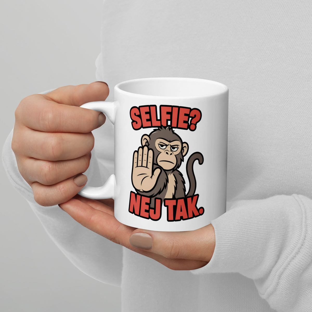 Hvidt keramikkrus med teksten Selfie? Nej tak og en abe der holder hånden op, holdt i hænderne, hank til venstre